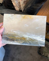 A hand holding a square ceramic tile or coaster featuring an abstract landscape painting with beige, white, gray, and gold brushstrokes.